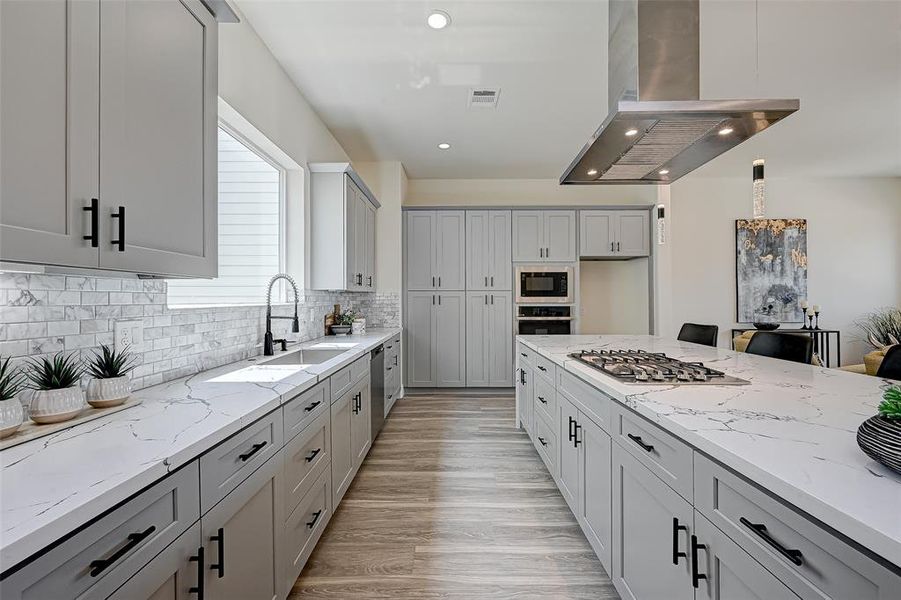 This modern kitchen features sleek gray cabinetry and white countertops. The space is brightened by a large window and accented with a stylish tile backsplash. An island with a built-in cooktop and overhead range hood offers ample workspace, while stainless steel appliances add a contemporary touch.