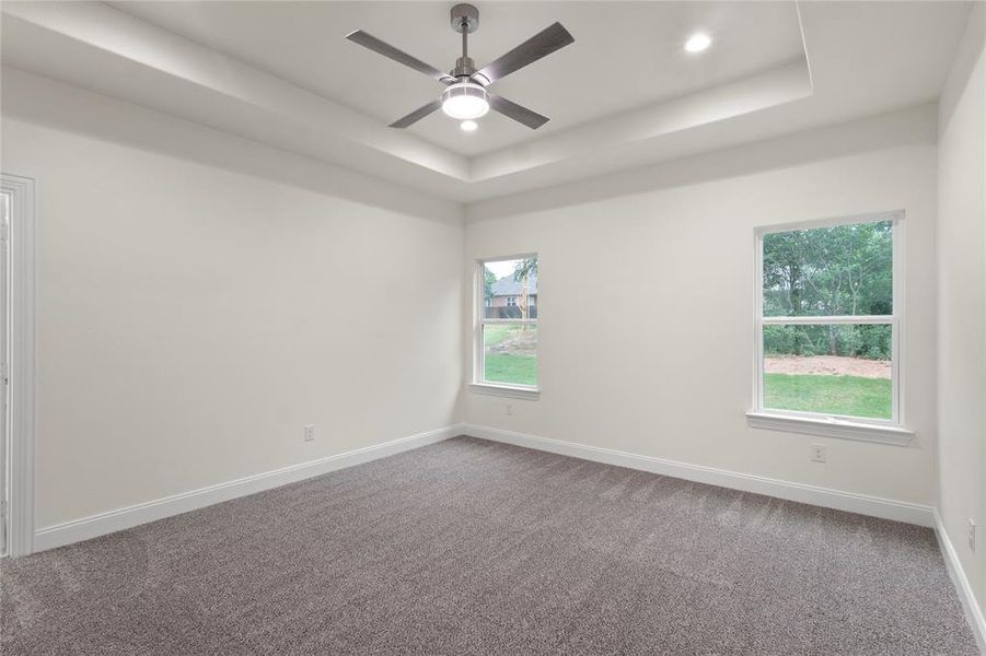 Spacious room featuring a tray ceiling with recessed lighting and a ceiling fan, plush gray carpeting, white baseboards, and two windows Spacious room featuring a tray ceiling with recessed lighting and a ceiling fan, plush gray carpeting, white baseboards, and two windows