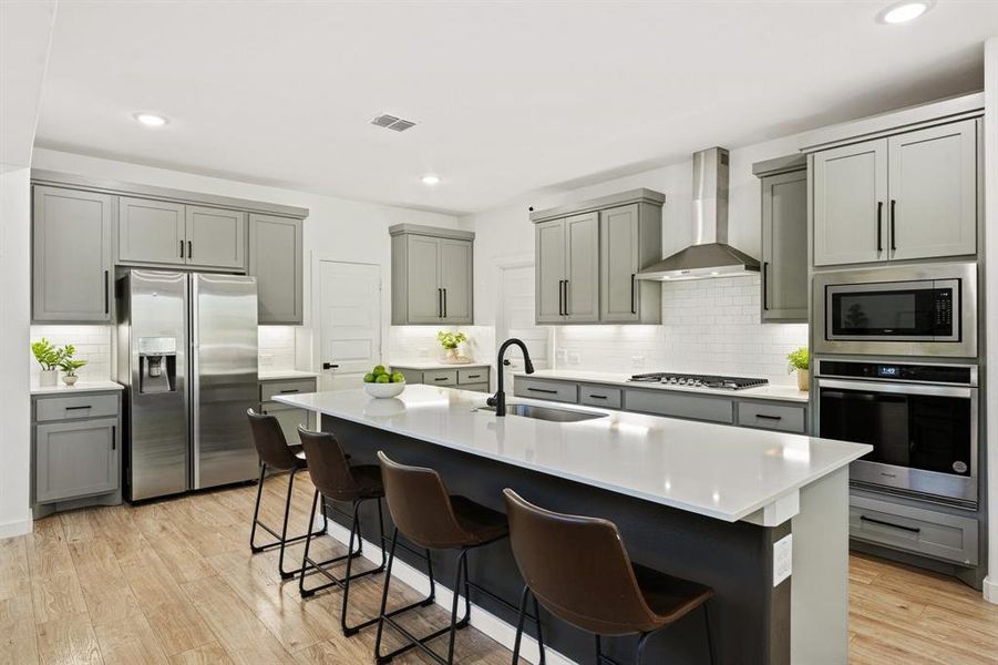 Kitchen featuring gray cabinetry, stainless steel appliances, a center island with sink, wall chimney exhaust hood, and a breakfast bar area