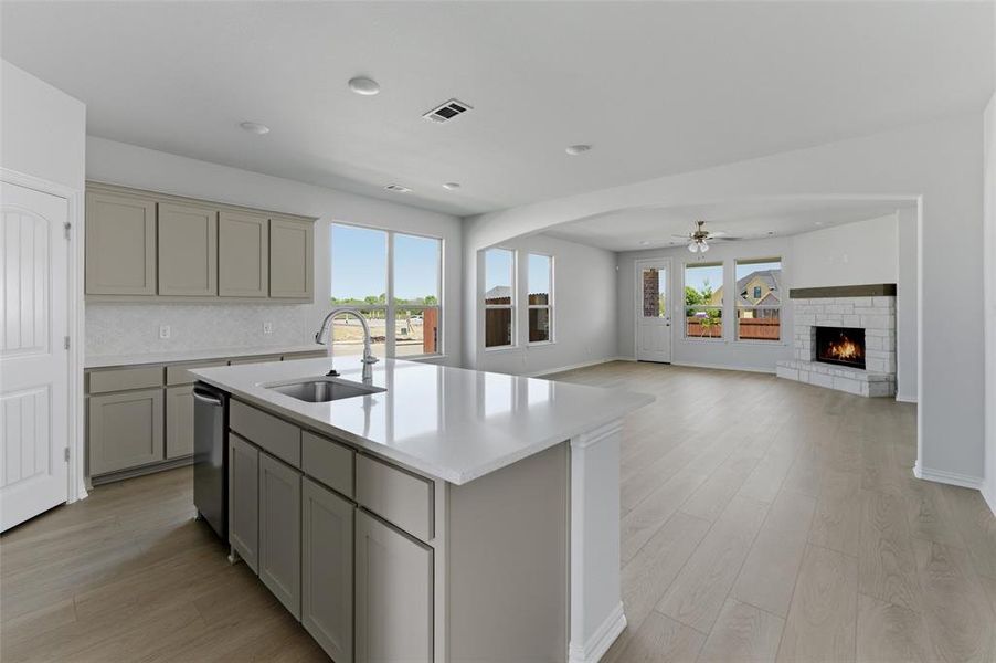 Kitchen featuring light wood-style floors, a warm lit fireplace, an island with sink, ceiling fan, and gray cabinets