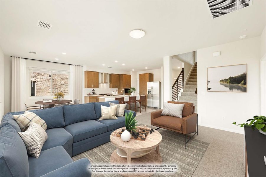 Open-concept living room with blue sectional sofa, connected kitchen with wood cabinets, and staircase visible in background. Open-concept living room with blue sectional sofa, connected kitchen with wood cabinets, and staircase visible in background.