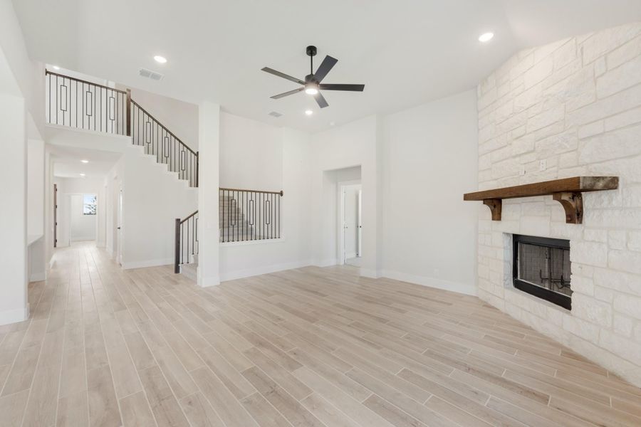Open living room with stone fireplace, wood mantel, light floors, ceiling fan, and staircase with iron railings