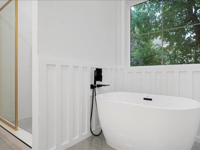 Bathroom featuring a freestanding bathtub with a wall-mounted faucet, a walk-in shower with a gold-framed glass enclosure, and a window with a view of greenery