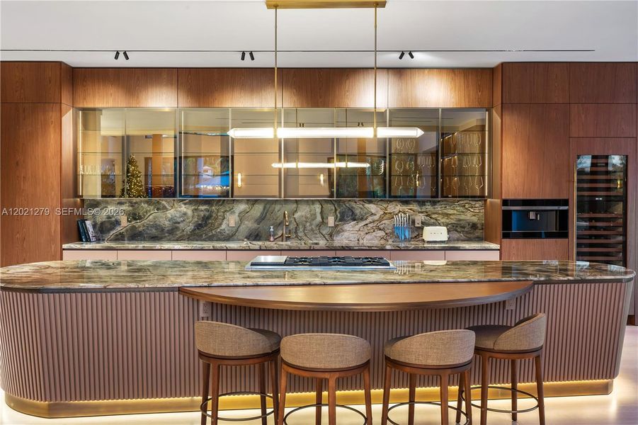 Gourmet kitchen featuring oversized curved island with book-matched stone countertop, fluted base, gas cooktop, bronze-framed glass upper cabinets, full-height walnut cabinetry, statement backsplash, wine refrigerator, built-in ovens, and brass linear pendant lighting.