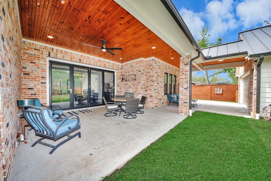 This photo showcases a spacious covered patio with a brick exterior, wooden ceiling, and ceiling fan. It features comfortable outdoor seating and a dining area, overlooking a well-maintained yard. Ideal for entertaining and relaxation.