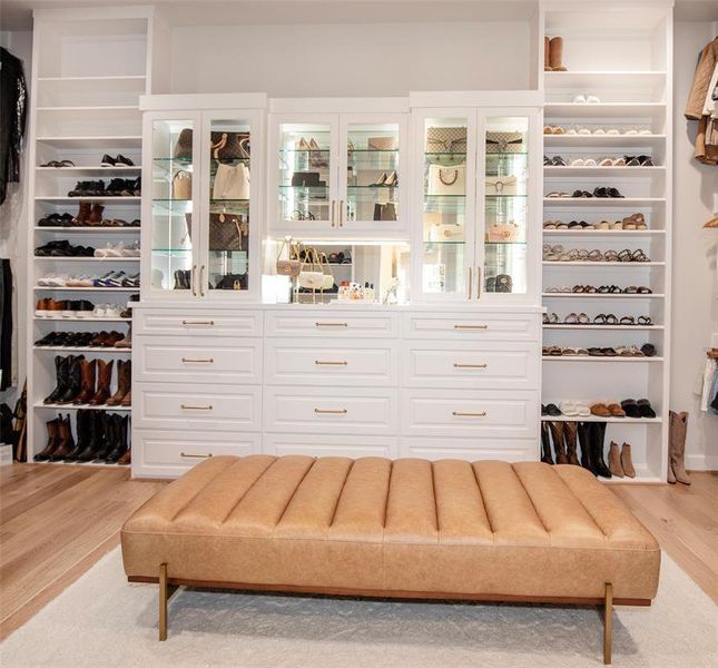 This photo showcases a spacious, well-organized walk-in closet with built-in shelves and cabinets for shoes and accessories. It features a chic, upholstered bench in the center and has a bright, elegant design with ample storage space.