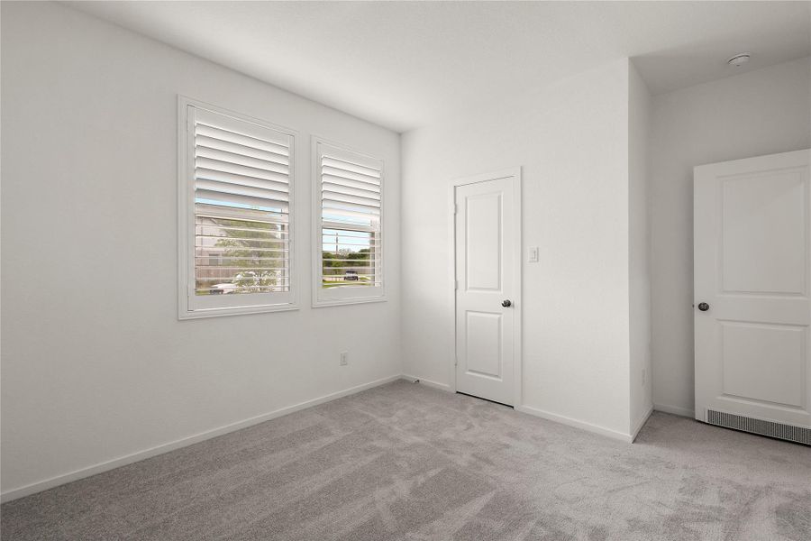 This room features soft gray carpeting, neutral white walls, and two windows with shutters, allowing natural light to brighten the space. It includes two doors, likely for a closet and entry, offering functionality and storage options.
