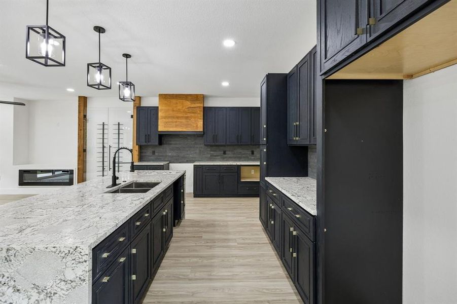 Spacious kitchen featuring a large island with an undermount sink, dark cabinetry, light-toned countertops, a full backsplash, and contemporary pendant lighting Spacious kitchen featuring a large island with an undermount sink, dark cabinetry, light-toned countertops, a full backsplash, and contemporary pendant lighting