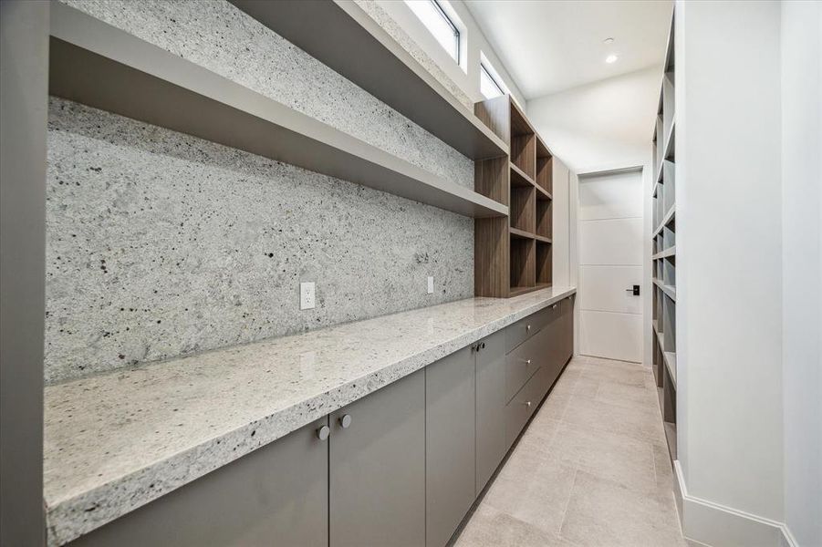 The pantry is outfitted with dune-carved natural floor tile and recessed LED lighting, enhancing its functionality and ambiance. Designer neutral wall paint complements the abundant built-in shelving, providing ample space for storage and organization.