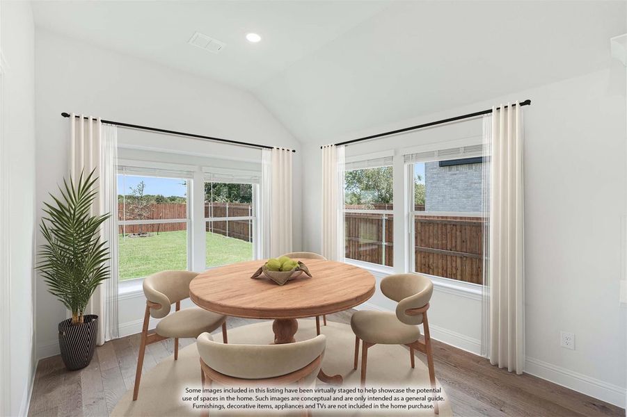 Bright dining area with round wood table, four chairs, vaulted ceiling, and large windows overlooking a fenced backyard.