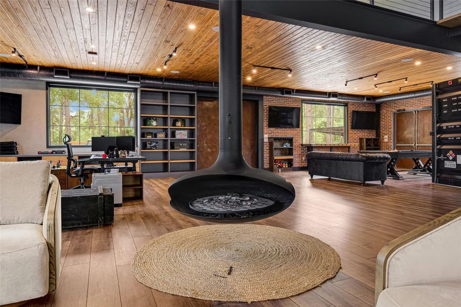 Suspended fireplace, warm wood flooring, and ceiling. Large windows offer natural light and views of the surrounding trees. The open layout includes a cozy seating area, a home office, and a recreation space.