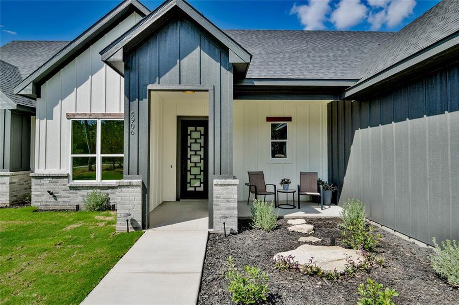 Doorway to property with board and batten siding, a shingled roof, brick siding, a porch, and a lawn Doorway to property with board and batten siding, a shingled roof, brick siding, a porch, and a lawn