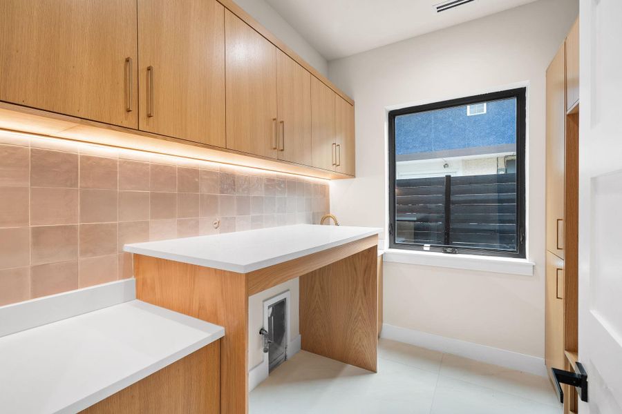 Laundry room featuring extensive light wood-finish cabinetry, white countertops, square tile backsplash with under-cabinet lighting, a single pane window, and light tile flooring