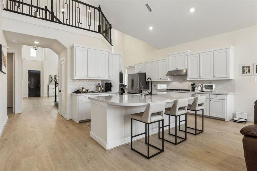 Open-concept kitchen with a large island, light wood-finish flooring, white cabinetry, a herringbone tile backsplash, and a stainless steel range hood