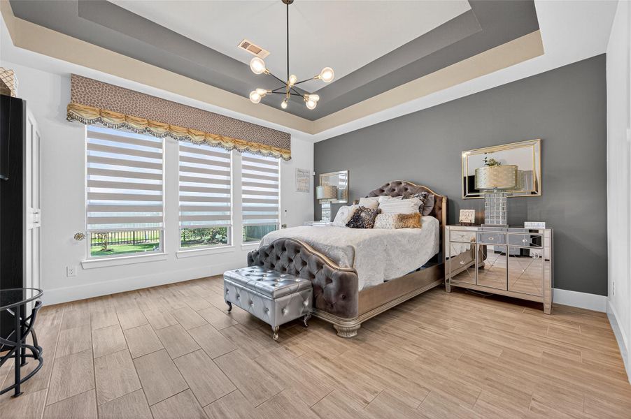This spacious bedroom features a modern design with a tray ceiling and stylish chandelier. It has large windows for natural light, a chic accent wall, and elegant furnishings, including a tufted bed and mirrored nightstands. The wood-look flooring adds warmth to the space. This spacious bedroom features a modern design with a tray ceiling and stylish chandelier. It has large windows for natural light, a chic accent wall, and elegant furnishings, including a tufted bed and mirrored nightstands. The wood-look flooring adds warmth to the space.