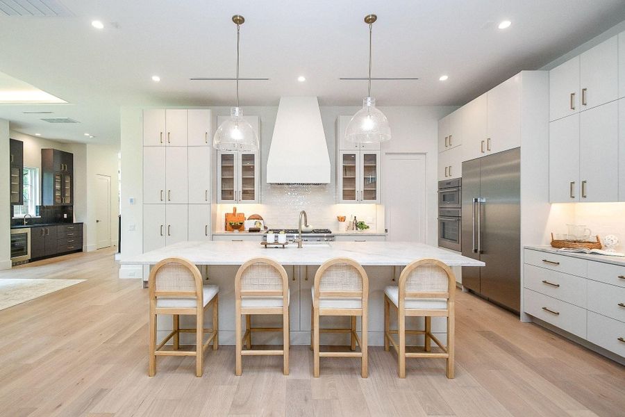 This kitchen features a spacious, modern design with a large island and seating for four. It has sleek white cabinetry, stainless steel appliances, and stylish pendant lighting, creating a bright and inviting space.