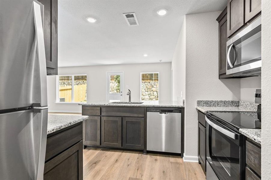 Kitchen with appliances with stainless steel finishes, a peninsula, dark brown cabinets, light wood-style floors, and light stone counters