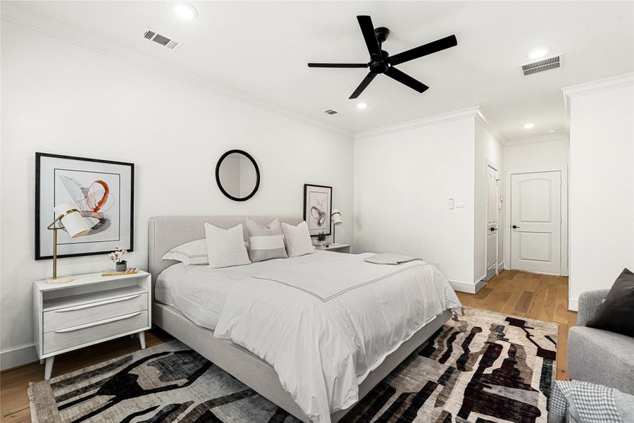 Modern, minimalist design with neutral tones. It includes a comfortable king-sized bed, and a ceiling fan. Natural light highlights the hardwood floors and ample recessed lighting.