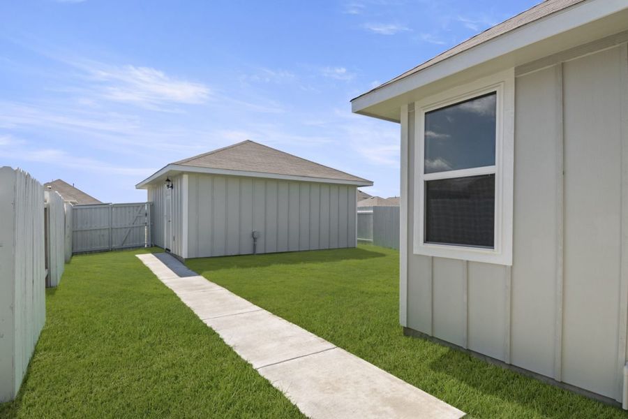 A backyard with a concrete walkway surrounded by grass leading to a gray building, and a white fence.