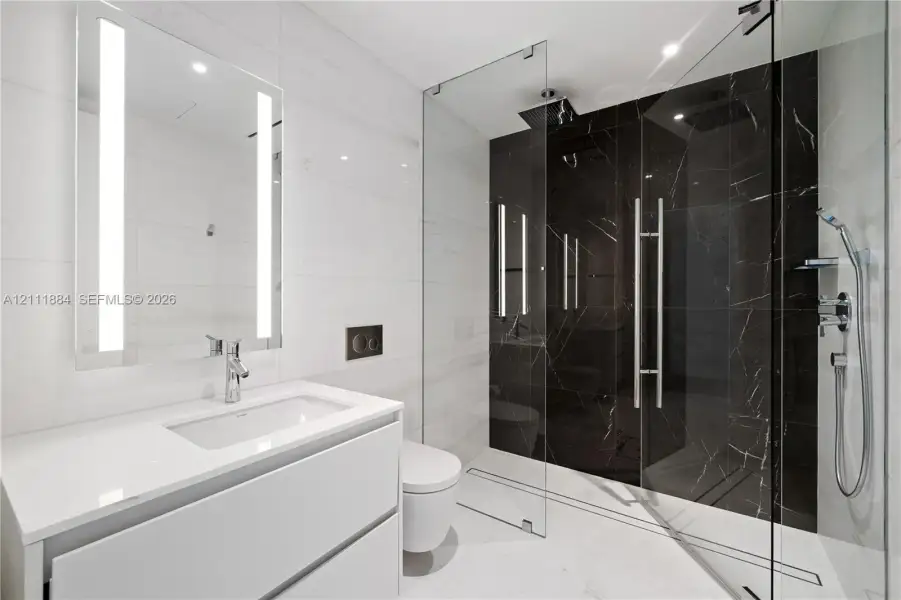Floating vanity with a white countertop and rectangular undermount sink, integrated LED-lit mirror, wall-mounted toilet with dual-flush actuator, and glass-enclosed shower with contrasting large-format tile surrounds and rainfall showerhead Floating vanity with a white countertop and rectangular undermount sink, integrated LED-lit mirror, wall-mounted toilet with dual-flush actuator, and glass-enclosed shower with contrasting large-format tile surrounds and rainfall showerhead
