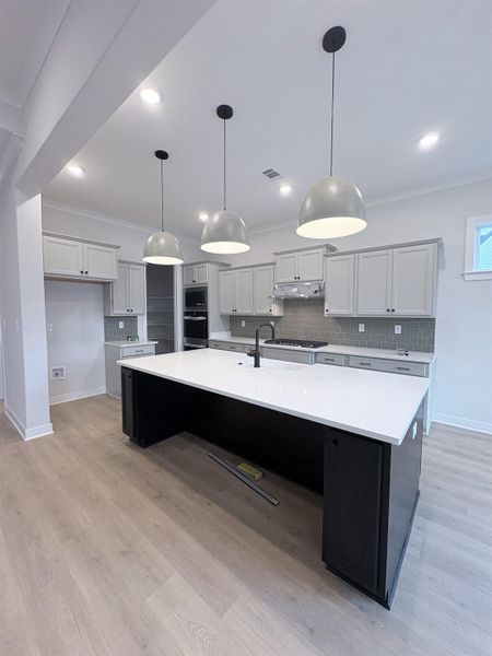 Contemporary kitchen featuring a large central island with a white countertop, light wood-finish flooring, grey cabinetry, a tile backsplash, and three pendant lights Contemporary kitchen featuring a large central island with a white countertop, light wood-finish flooring, grey cabinetry, a tile backsplash, and three pendant lights