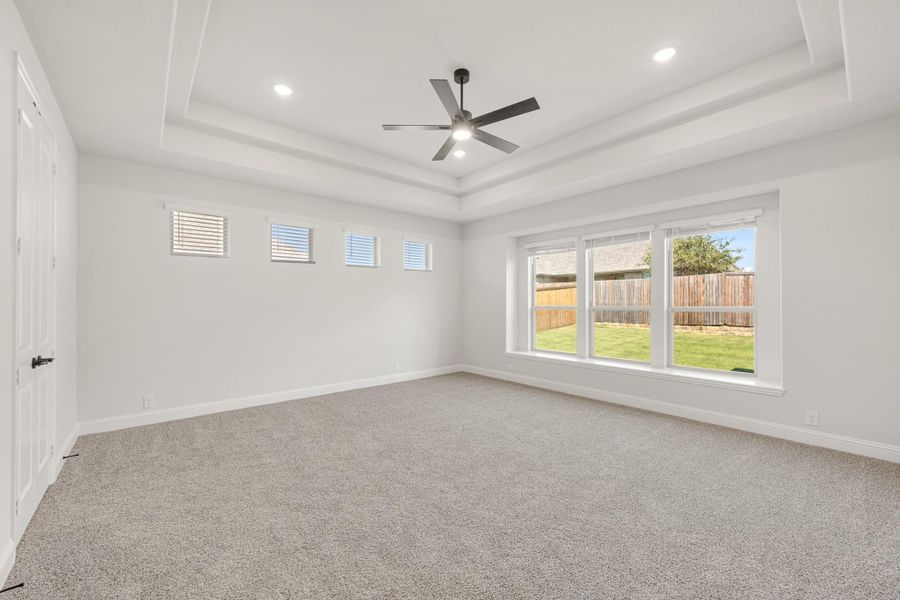 Empty bedroom with tray ceiling, ceiling fan, carpet flooring, and large window overlooking backyard Empty bedroom with tray ceiling, ceiling fan, carpet flooring, and large window overlooking backyard