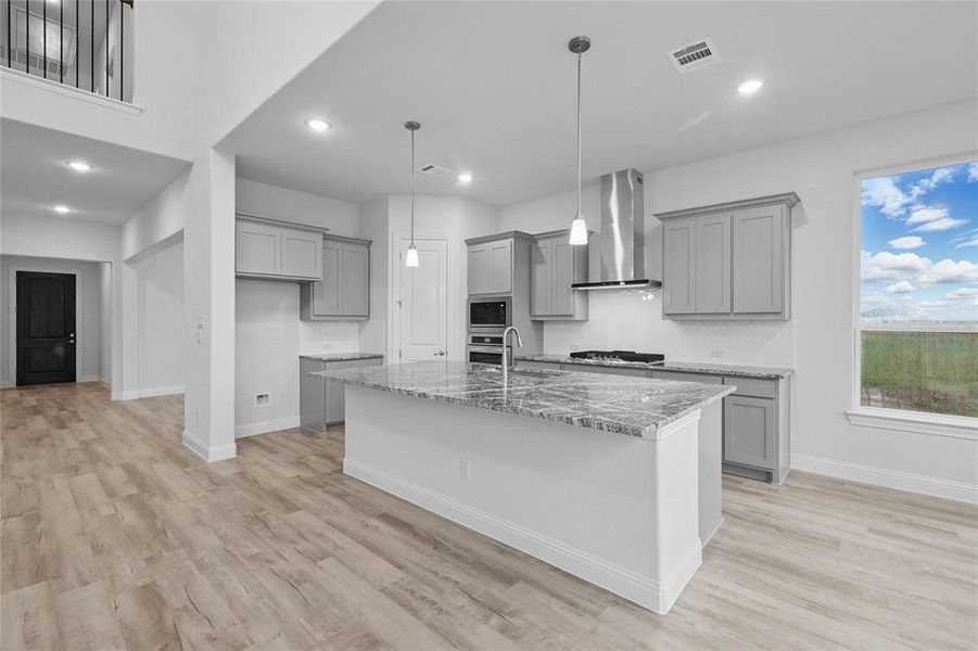 Kitchen with gray cabinetry, wall chimney range hood, light stone counters, appliances with stainless steel finishes, and baseboards