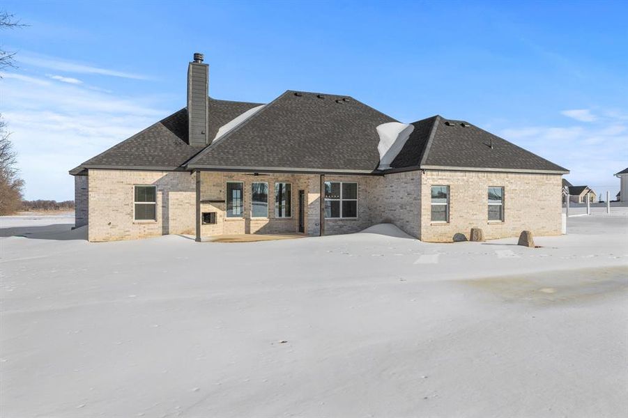 Snow covered back of property featuring brick siding, a chimney, a patio, and a shingled roof