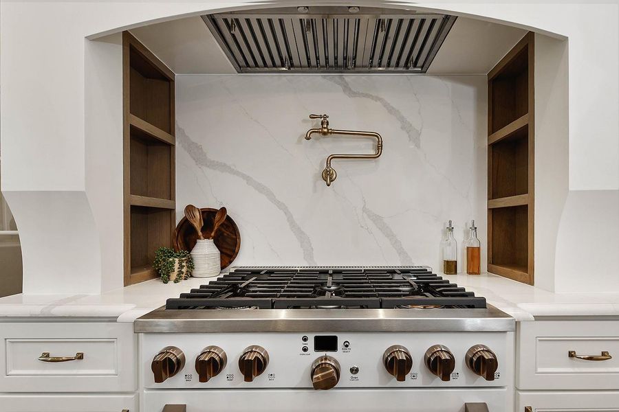 Custom cooking section has enclosed vent hood, and hidden spice shelves flanking a premium GE Cafe gas range cooktop and oven in white with gold knobs. Convenient gold pot filler is upscale and a highly desirable feature.