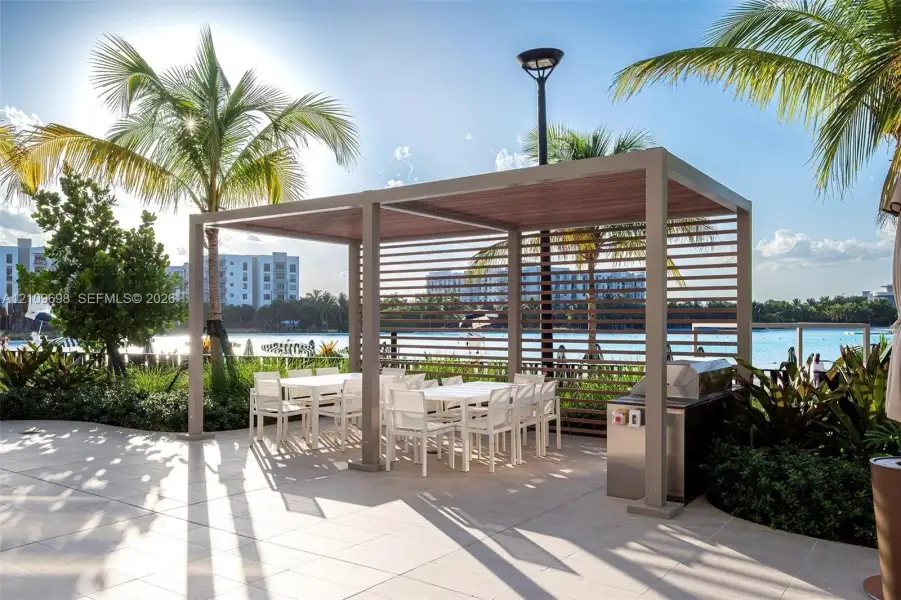 Outdoor patio featuring a rectangular metal-framed pergola with slatted walls, integrated stainless steel grill, large-format tile flooring, and mature palm trees