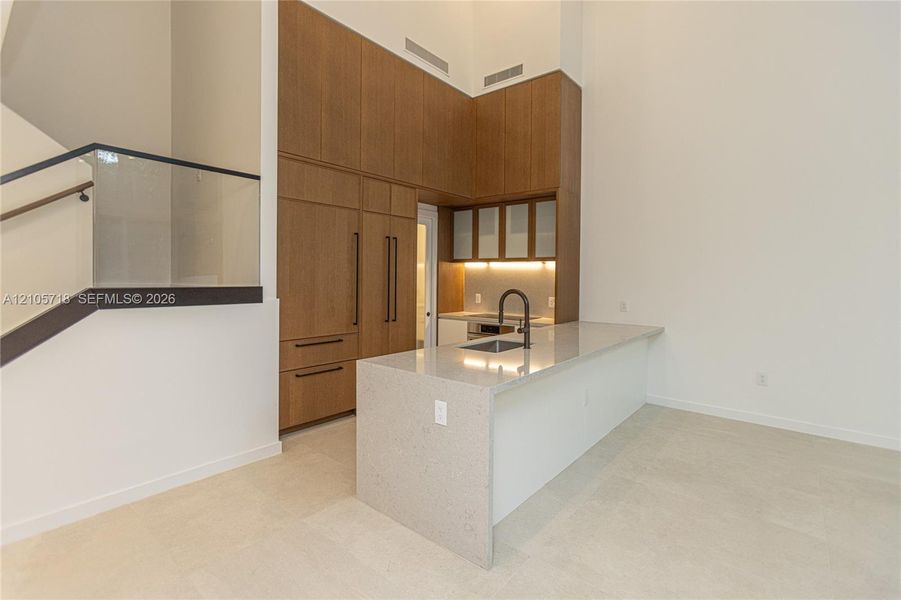 Kitchen featuring full-height wood-finish cabinetry, waterfall-edge stone countertop, undermount sink with matte black faucet, and glass staircase railing