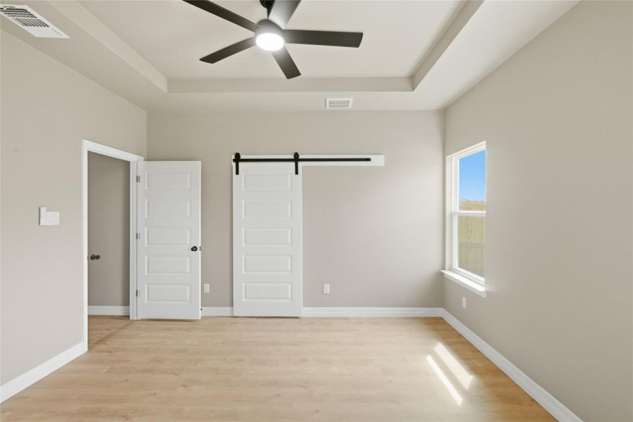 Unfurnished bedroom featuring a barn door, a raised ceiling, light wood finished floors, and a ceiling fan
