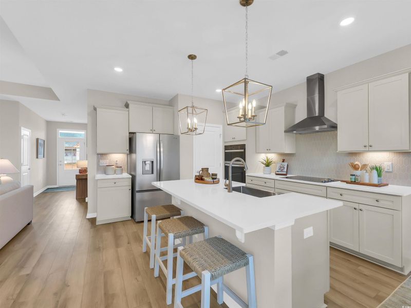 Kitchen with backsplash, stainless steel refrigerator with ice dispenser, wall chimney range hood, hanging light fixtures, and light wood-style floors