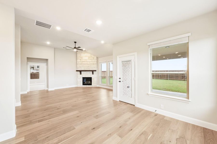 Open living room with light hardwood floors, stone fireplace, ceiling fan, and backyard views through large windows
