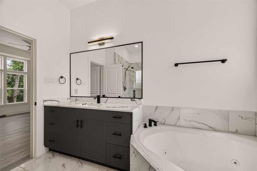 This modern bathroom features a spacious vanity with dual sinks, a large mirror, and sleek black fixtures. The corner bathtub is surrounded by elegant marble tile, and there's a glimpse of a bright room with large windows through the open door. This modern bathroom features a spacious vanity with dual sinks, a large mirror, and sleek black fixtures. The corner bathtub is surrounded by elegant marble tile, and there's a glimpse of a bright room with large windows through the open door.