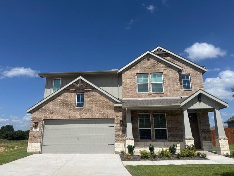 Two-story residence featuring a brick and stone facade with vertical siding accents, an attached two-car garage, covered entry porch, and mature landscaping Two-story residence featuring a brick and stone facade with vertical siding accents, an attached two-car garage, covered entry porch, and mature landscaping