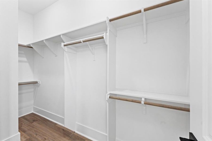 This photo shows a spacious, walk-in closet with multiple wooden hanging rods and white shelving, perfect for organizing clothes and accessories. The space is bright and has a clean, modern look with light wood flooring.