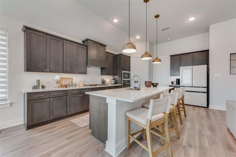 Kitchen featuring a central island with integrated sink, dark wood cabinetry, white countertops, coordinating white subway tile backsplash, and light wood-finish flooring