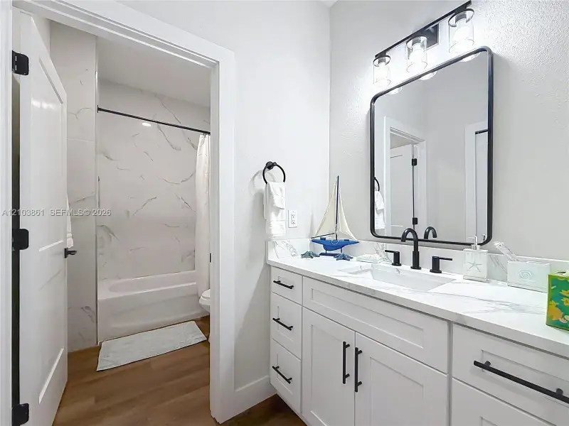 Bathroom featuring a white vanity with stone-style countertop, rectangular undermount sink, matte black hardware, framed vanity mirror, and tub-shower combination with floor-to-ceiling tile surround