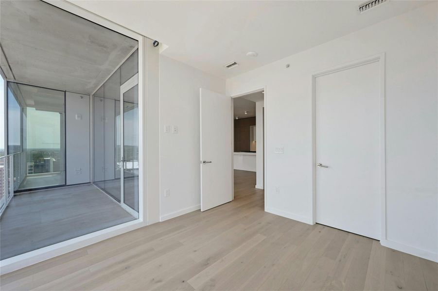Bright and airy bedroom showcasing expansive floor-to-ceiling windows that frame stunning city views. Light wood flooring and clean, modern finishes create a sleek and tranquil retreat. The room offers easy access to the main living area and includes a spacious closet for added functionality.