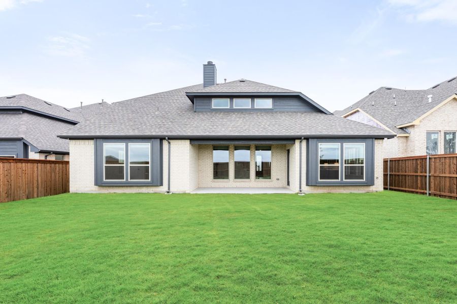 Rear view of single-story brick home with large windows, covered patio, and fenced green backyard