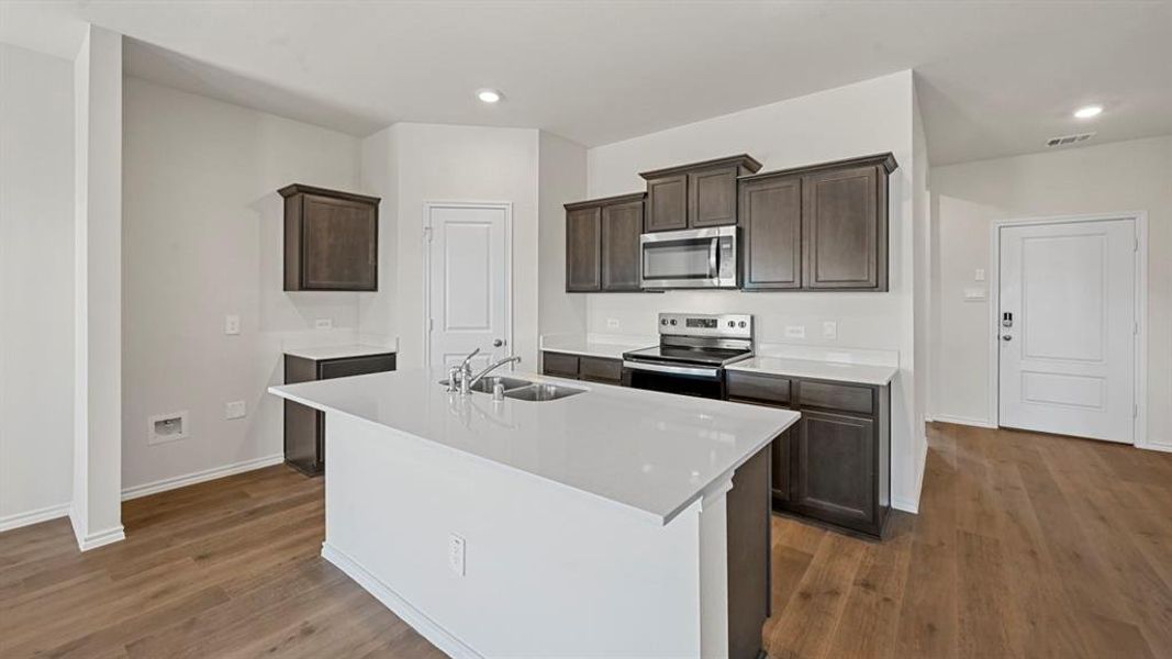 Modern kitchen featuring a central island with an undermount double sink, white countertops, dark wood-finish cabinetry, stainless steel appliances, and wood-finish flooring