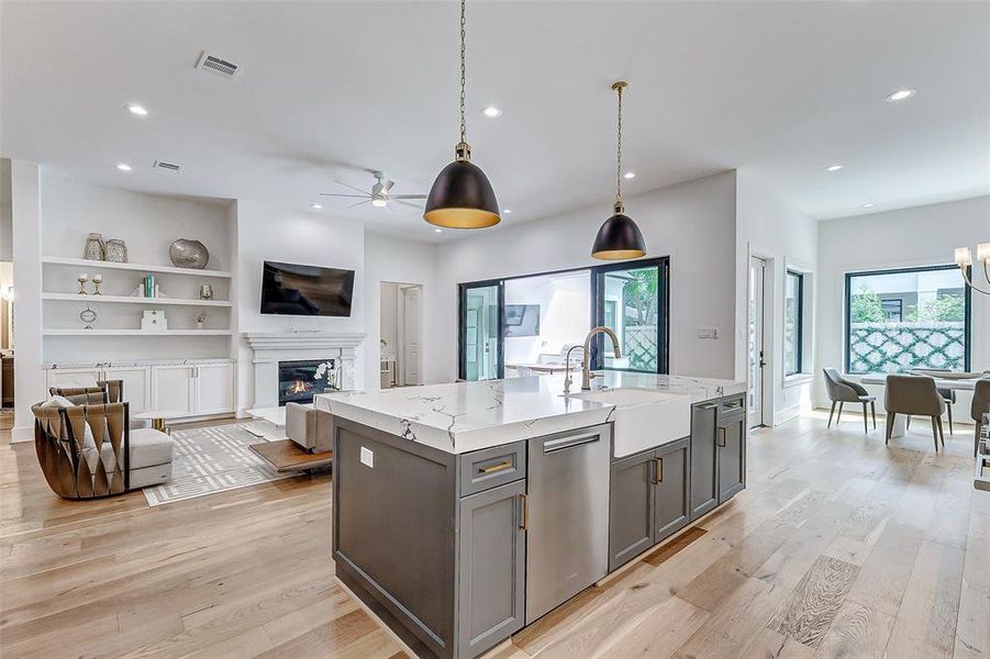 This modern open-concept living space features a bright and airy design, with hardwood floors, a large kitchen island with a farmhouse sink, a Miele dishwasher, and stylish pendant lighting. The living area includes a cozy fireplace and built-in shelving, while large windows offer plenty of natural light.