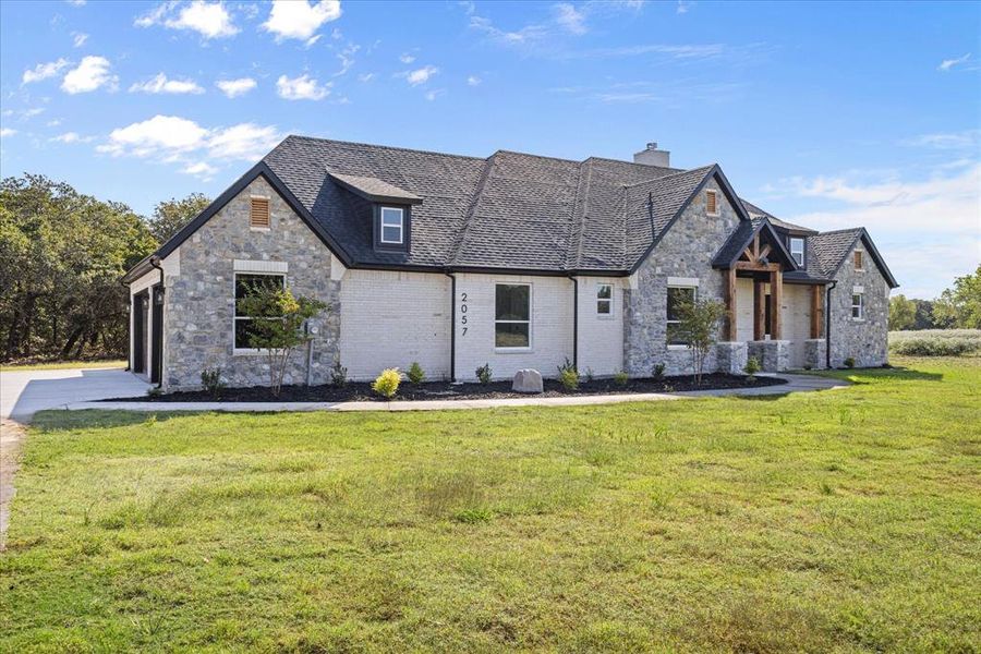 Modern farmhouse exterior featuring a stone and white brick facade, black-trimmed windows, a dark shingle roof, a covered entry with timber accents, and an attached two-car garage