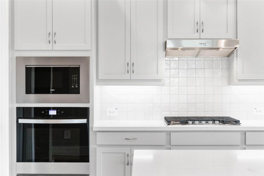 Top-of-the-line stainless steel appliances that offer both professional function and modern style. The focal point is a professional-grade gas cooktop with multiple burners and robust grates, perfectly complemented by an elegant stainless steel range hood. A built-in microwave conveniently located above the coordinating built-in oven, creating a highly efficient and visually appealing cooking center that streamlines meal preparation.