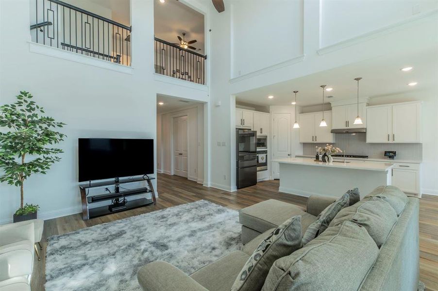 Open-concept living space featuring wood-finish flooring, a kitchen with white cabinetry, a central island with countertop seating, and a double-height ceiling