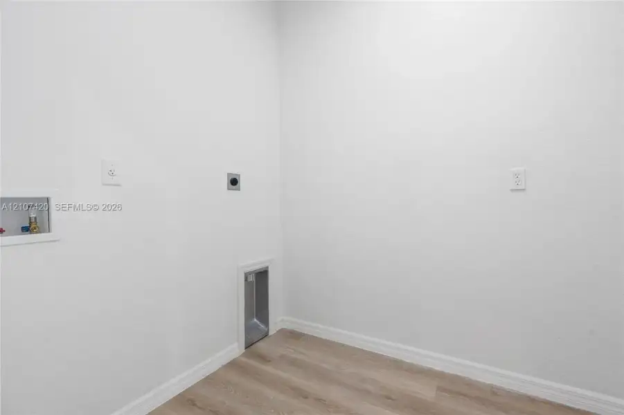 Utility area featuring wood-finish flooring, recessed water supply and drainage hookups, and a dedicated wall-mounted dryer vent Utility area featuring wood-finish flooring, recessed water supply and drainage hookups, and a dedicated wall-mounted dryer vent