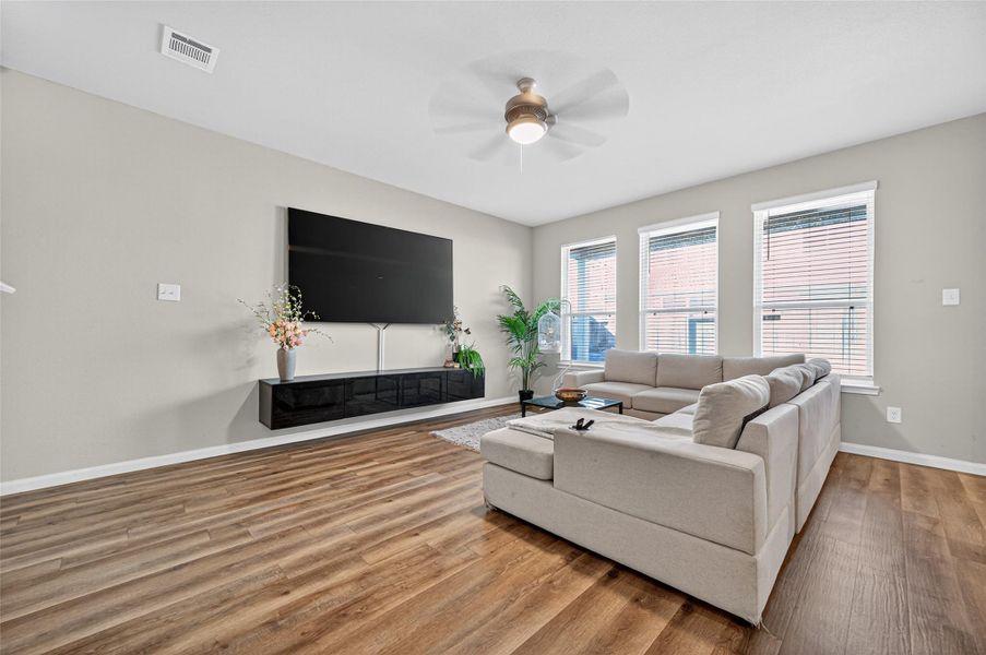 Spacious living room with modern wood flooring, large sectional sofa, and a wall-mounted TV. Ample natural light streams through three windows, complemented by a ceiling fan and tasteful decor.