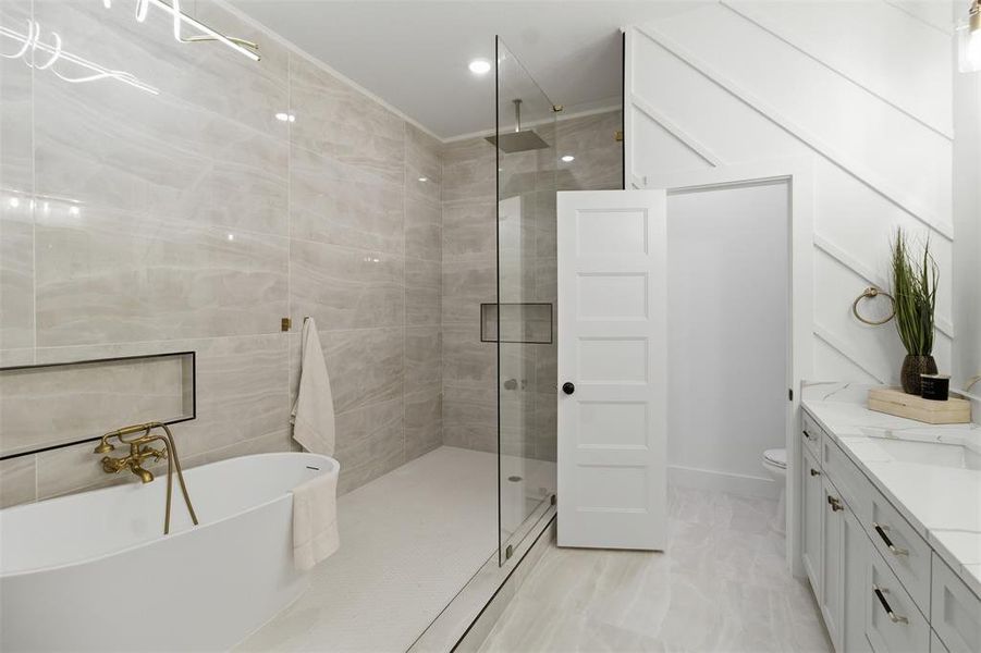 Spacious bathroom featuring a freestanding soaking tub with brass fixtures, a glass-enclosed shower with tiled walls and a recessed niche, a double vanity with a marble-finish countertop, and a decorative accent wall