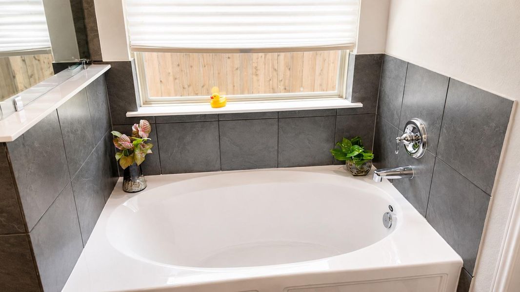 This bathroom features a white soaking tub with a chrome faucet and a dark gray tile surround. A single window with a horizontal privacy shade is positioned directly above the tub.